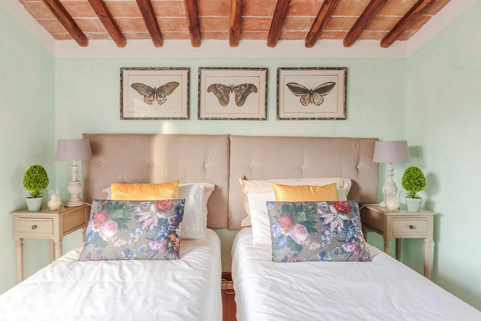 Una cama doble con almohadas estampadas, un cabecero color beige y dos mesitas de noche con lámparas y pequeñas macetas.