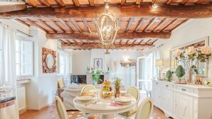 A dining area with a round table set for four, surrounded by chairs. A chandelier hangs from the ceiling, and the room features exposed wooden beams.