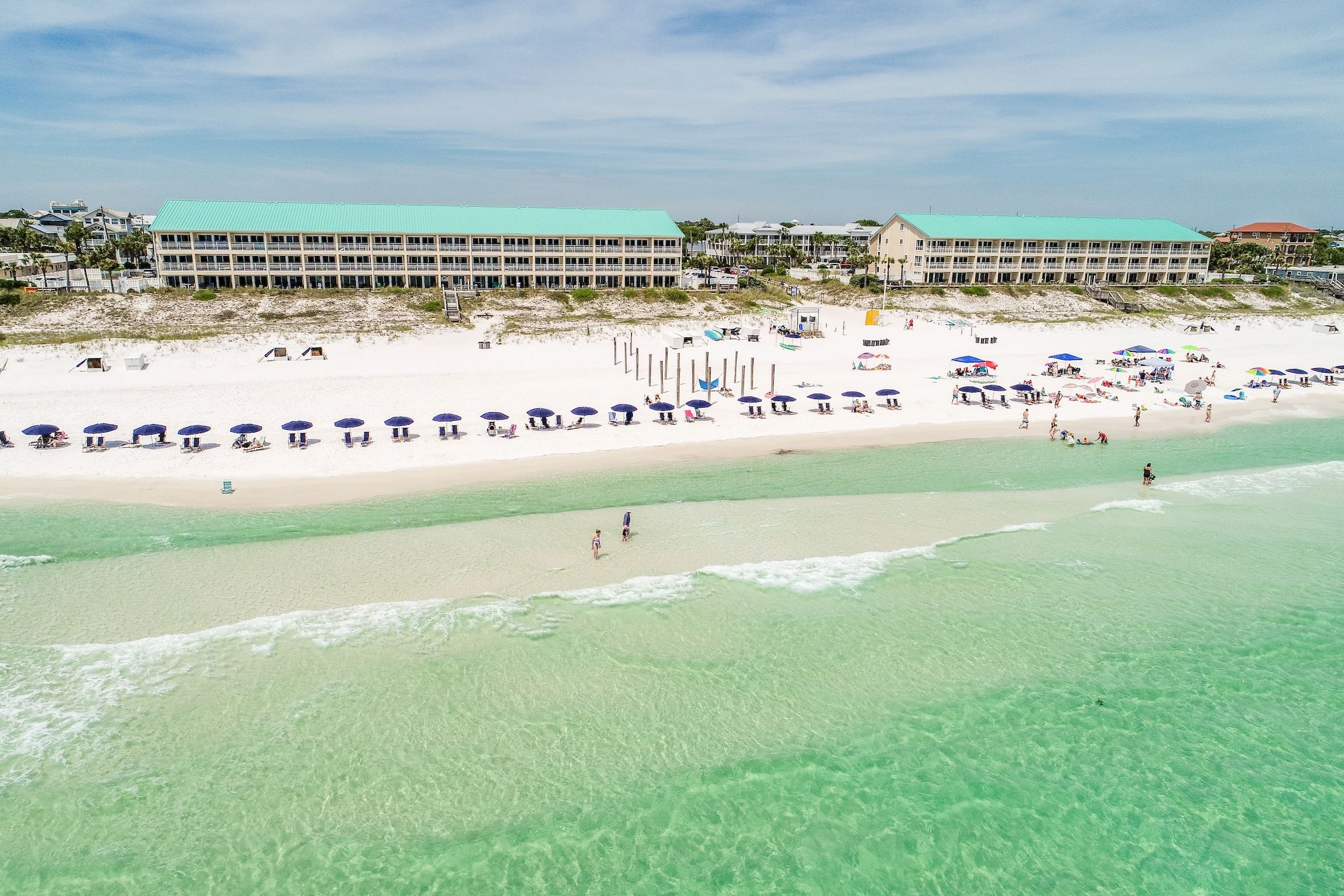 A beach with a row of beach umbrellas, a building with a green roof, and a clear view of the ocean.