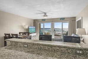 A room with a kitchen area, dining table, chairs, a TV, and a view of the ocean through large windows.