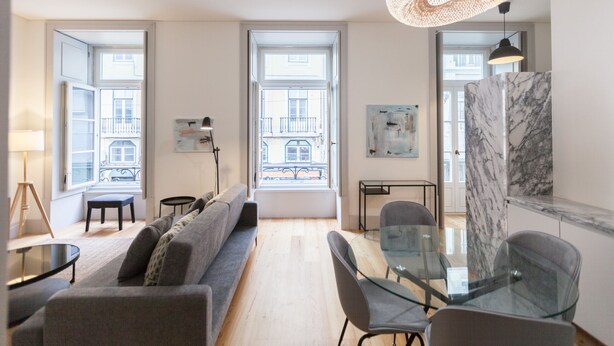 Ein modernes Wohnzimmer mit einer Couch, Glastisch und Stühlen, großen Fenstern mit Blick auf Gebäude und einer Hängeleuchte.
