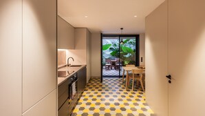 A compact kitchen with a modern design, featuring a sink, stove, and cabinets. The floor has a geometric pattern, and there is a dining area with a table and chairs visible through a glass door.