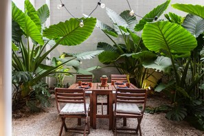 A wooden table with four chairs, surrounded by large green plants and a white tiled wall.
