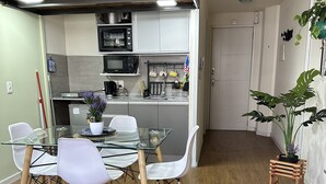 Una cocina compacta con electrodomésticos modernos, un comedor con mesa de vidrio y sillas blancas, y un taburete de madera con una planta.