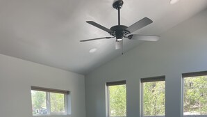 A spacious living area with a dark-colored sofa, a wooden coffee table, and a ceiling fan. There are large windows providing natural light and views of greenery outside.