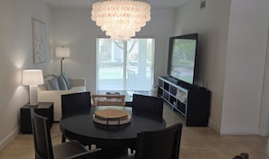 A dining area with a round table, chairs, a chandelier, a sofa, a TV, and a sliding glass door.