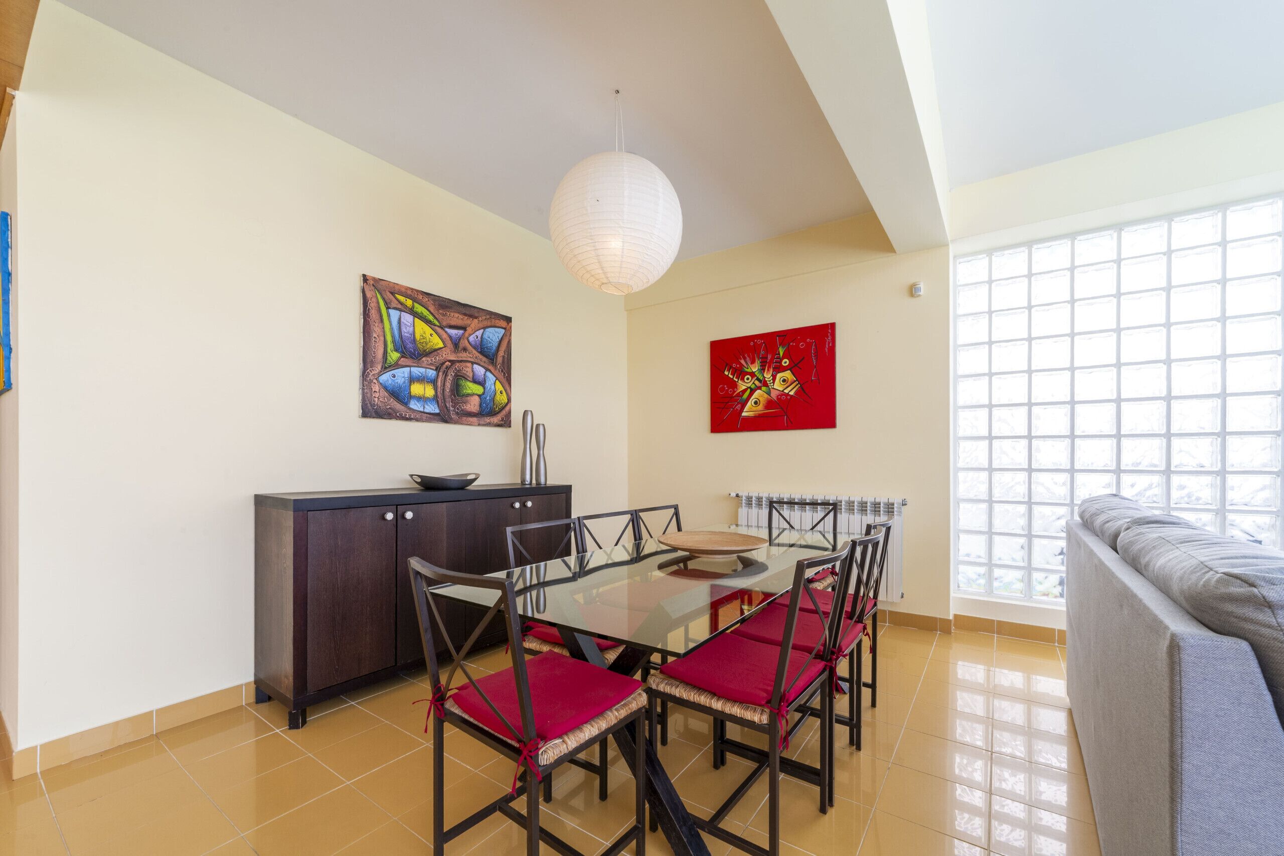 Un comedor con una mesa de vidrio, sillas rojas acolchadas, un aparador de madera oscura y dos cuadros colgados en la pared.