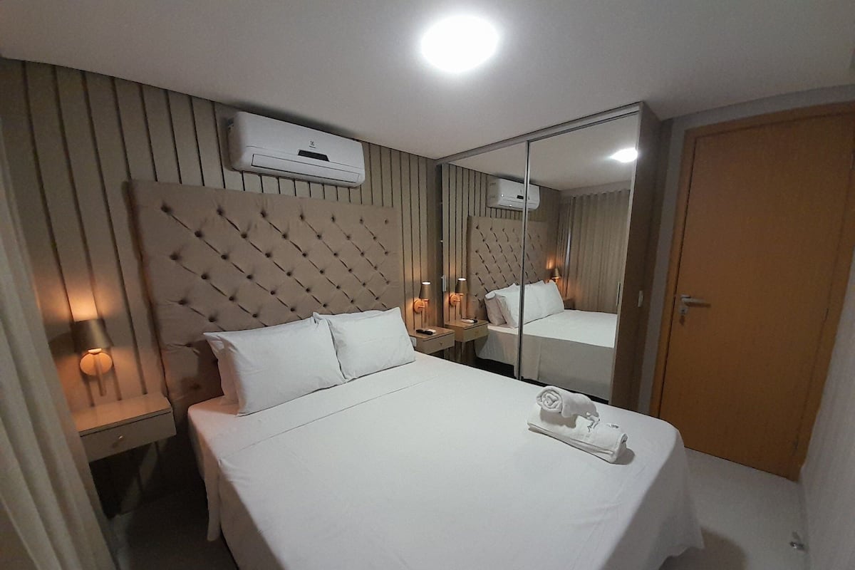 Ein Hotelzimmer mit einem Bett, einem Nachttisch, einer Lampe, einem Spiegel und einer Wand-Klimaanlage.