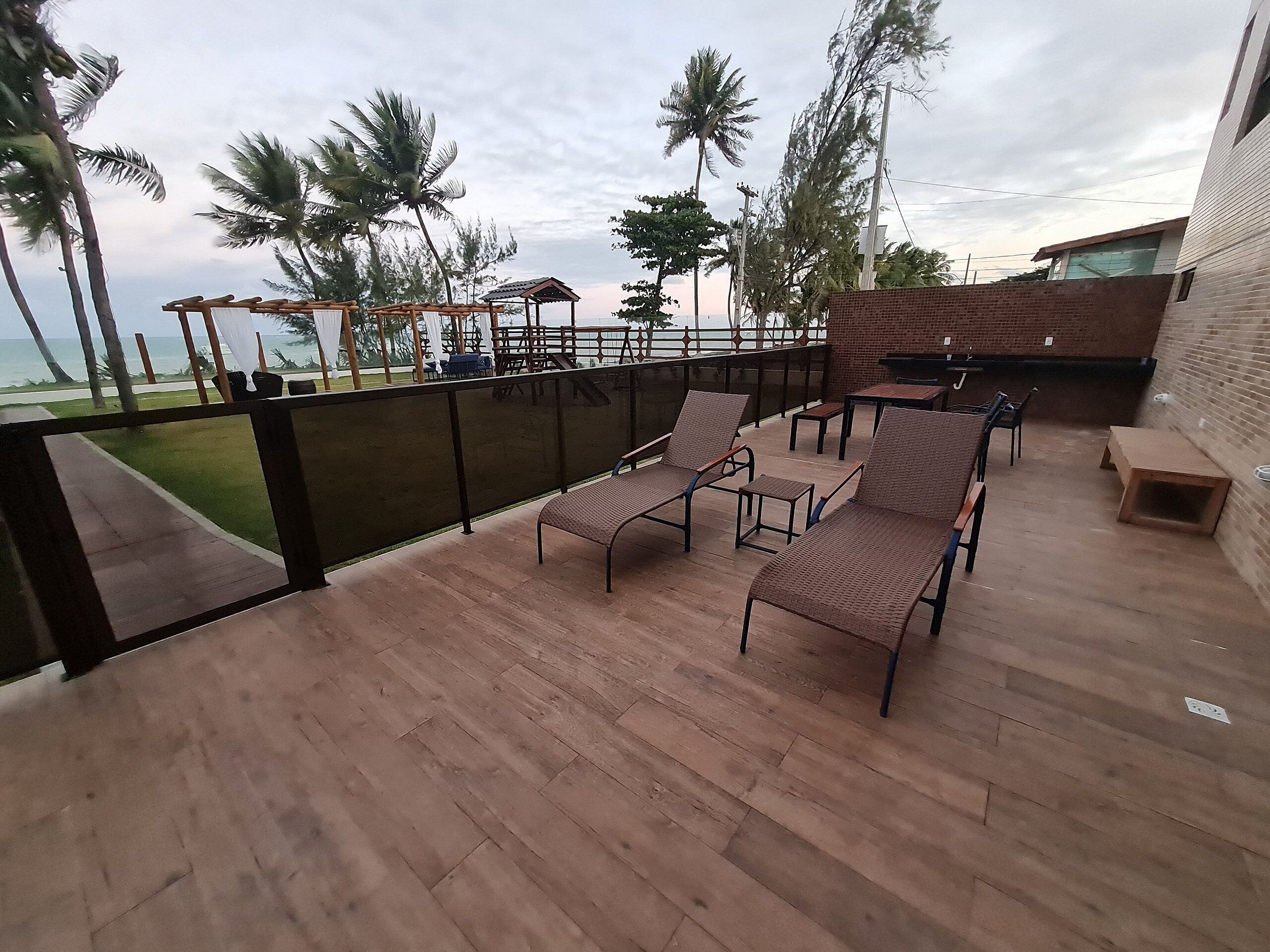Terraza de madera con sillones de descanso y un muro de ladrillo, con vista a una playa con palmeras.