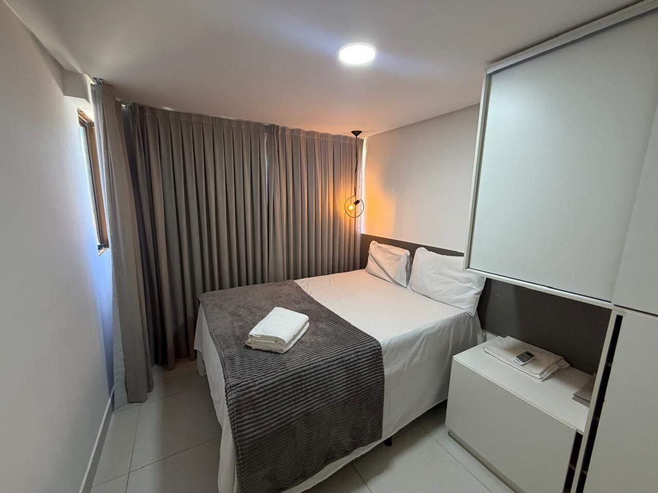 Een hotelkamer met een bed, nachtkastje, kledingkast en raam met gordijnen.