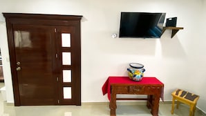 Ein Zimmer mit Holztür, einem an der Wand befestigten Fernseher, einem kleinen Tisch mit rotem Tischläufer und einer bunten Vase auf dem Tisch.