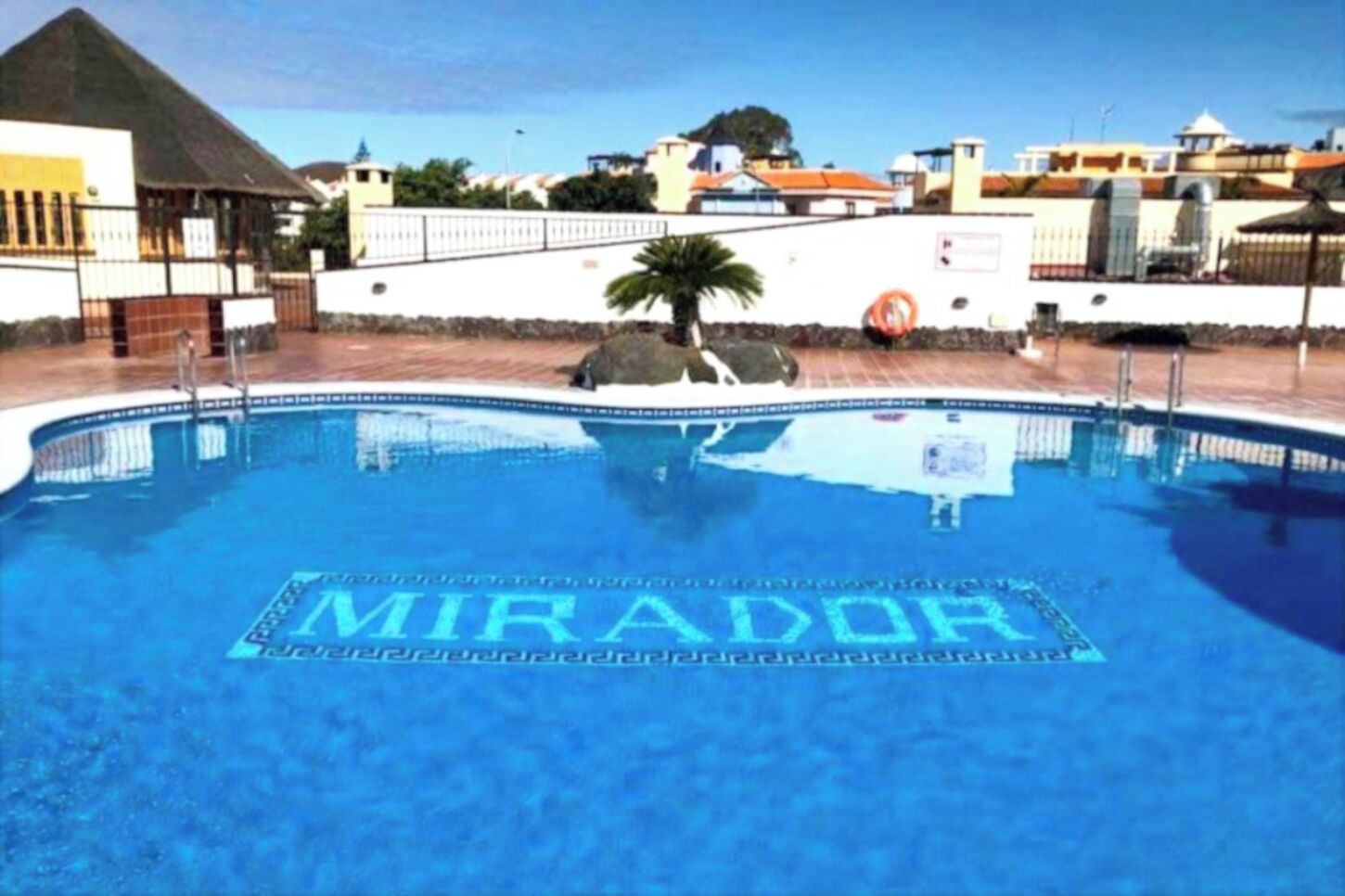 A swimming pool with the word "MIRADOR" on the water's surface, surrounded by a white wall and a palm tree.