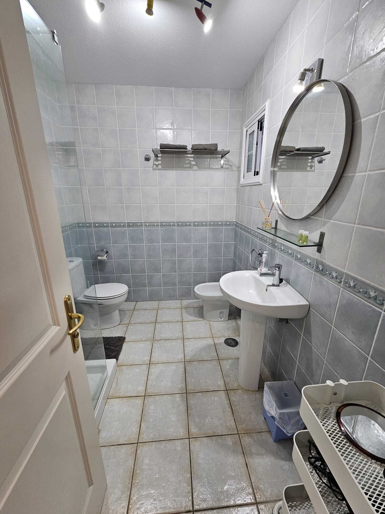 Een badkamer met twee wastafels, een toilet en een bidet. Er zijn planken met toiletartikelen en een spiegel aan de muur.
