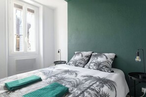Une chambre à coucher avec un lit, deux oreillers à motifs de palmiers, un mur vert, une fenêtre et une table de chevet avec une lampe.