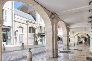 Une zone piétonne abritée, avec des arcades en pierre, un sol carrelé et un bâtiment doté d’une fenêtre grillagée et d’un pot de fleurs.