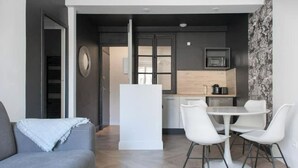 A modern living room with a grey sofa, a dining table with white chairs, a kitchen area with a microwave and wooden cabinets, and a patterned wall. - The Parrot 🦜 from La Rochelle 🐝 (La Rochelle)
