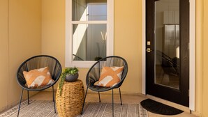 A corner with two wicker chairs with cushions, a small round wicker table with a plant, and a dark wooden door.