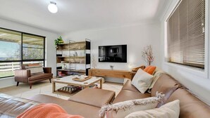 A modern living room with a sofa, coffee table, and a TV. There is a bookshelf and a window with blinds.