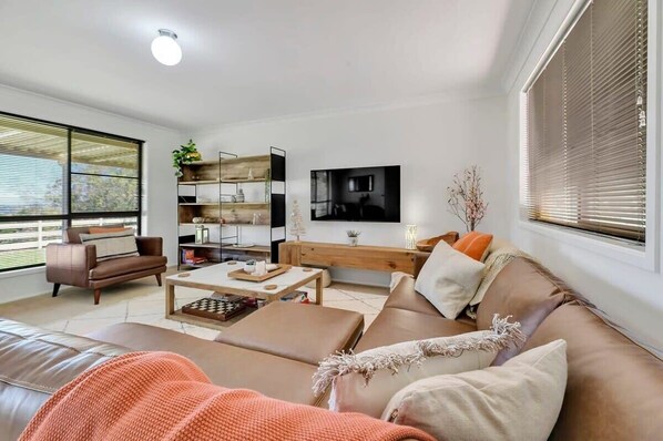 A modern living room with a sofa, coffee table, and a TV. There is a bookshelf and a window with blinds.