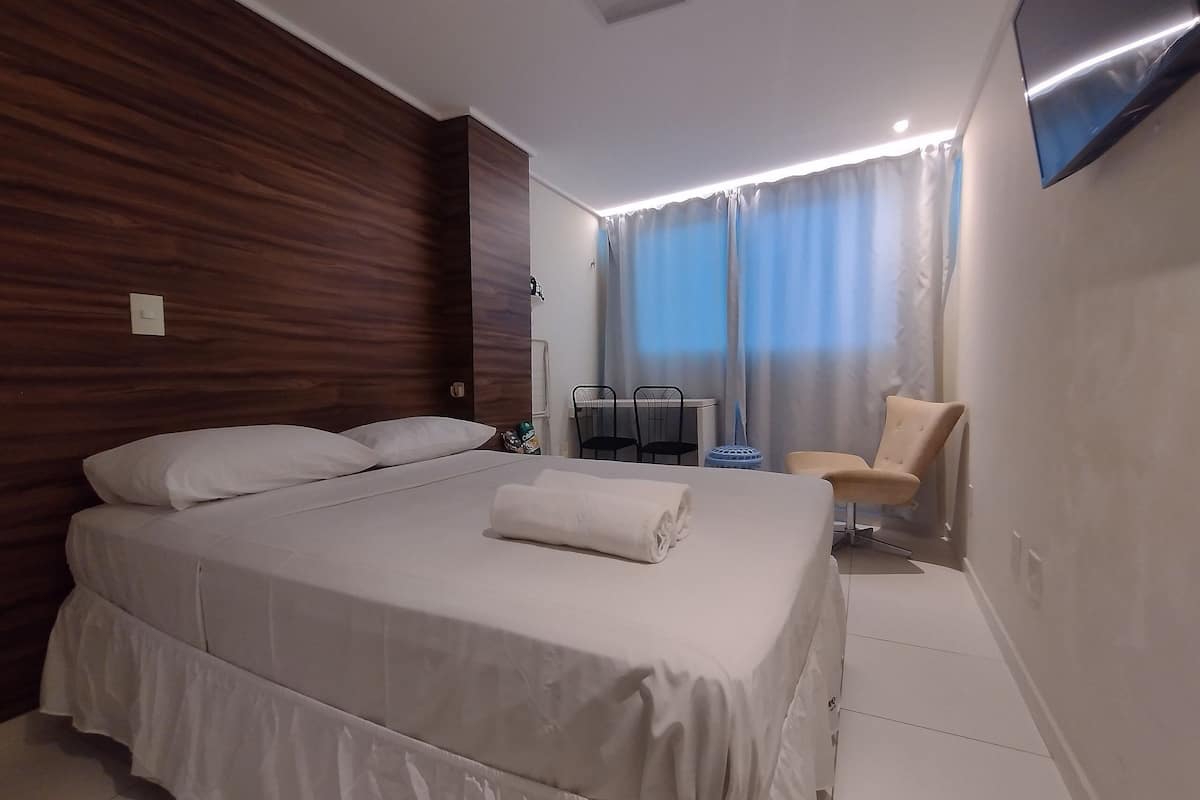 Quarto de hotel com cama, poltrona, televisão e janela com cortinas.