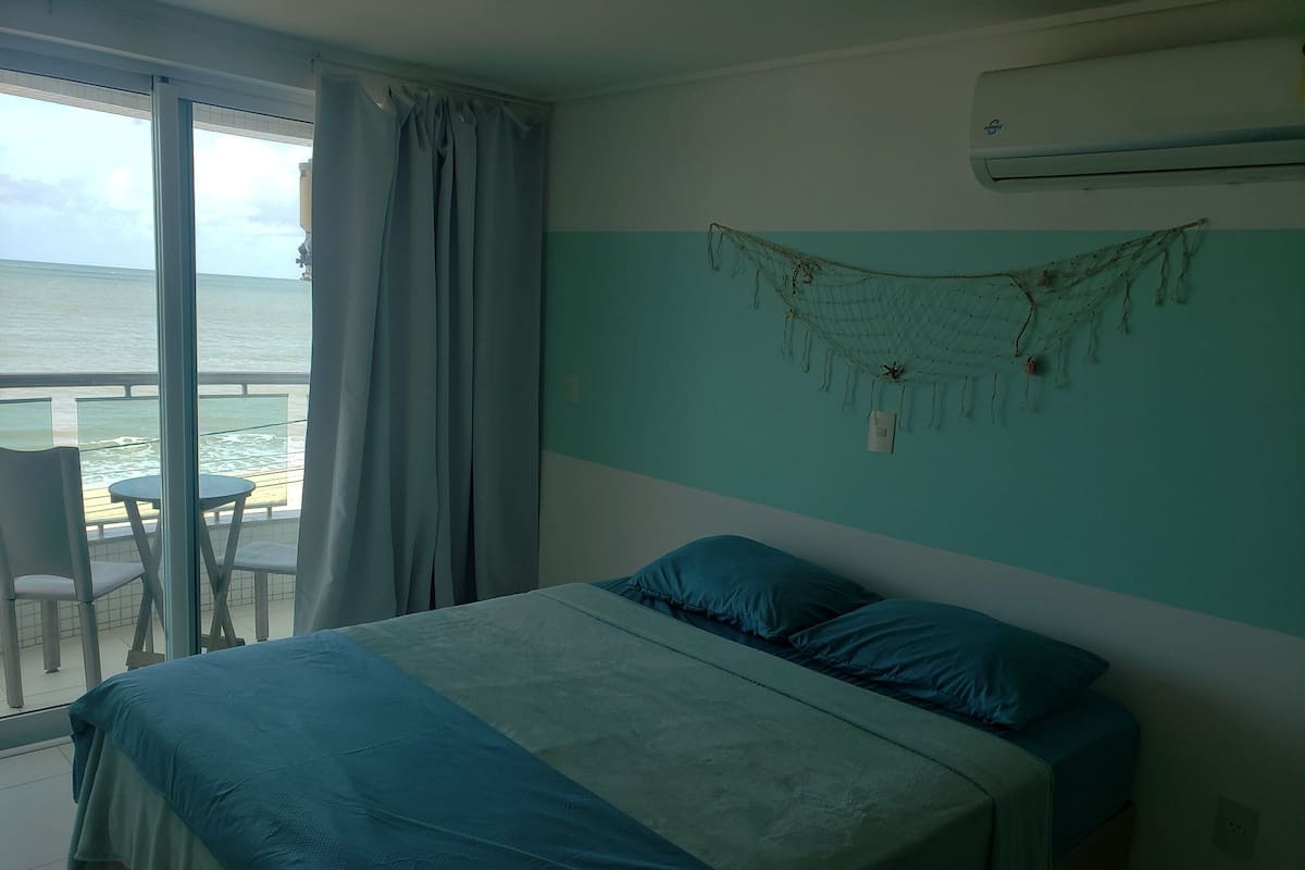 Quarto com uma cama, uma cadeira, uma mesa pequena e vista para o oceano.