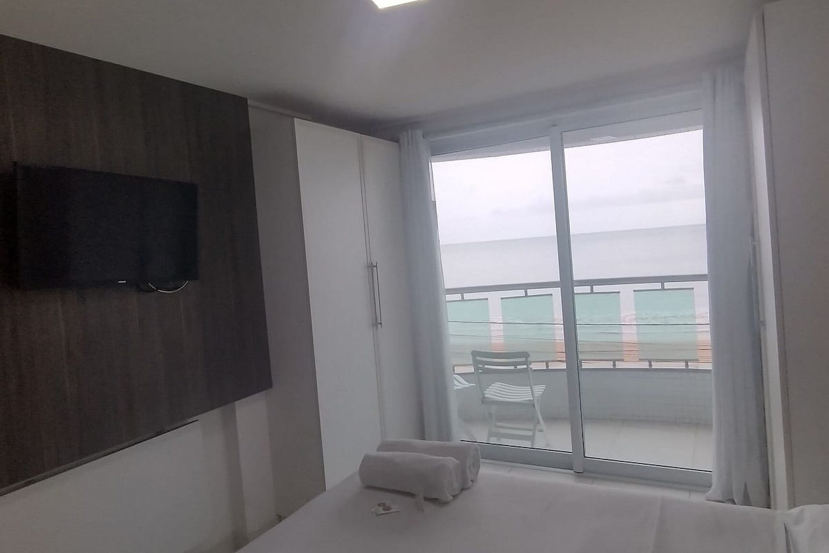 Quarto de hotel com cama, televisão na parede, janela vista para o mar e varanda com cadeira.