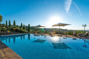 Una piscina azul cristalina con bordes de azulejos, rodeada de sillones de descanso y sombrillas, con un fondo de árboles y cielo despejado.