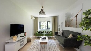 Ein modernes Wohnzimmer mit Fernseher, einem grünen Sofa, einem grauen Sofa, einem Couchtisch und einem gemusterten Teppich.
