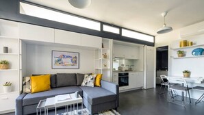 Een moderne woonkamer met een grijze bank, een bijzettafeltje en een kamerbreed tapijt. Er is een keuken met witte kasten en een eettafel met stoelen.