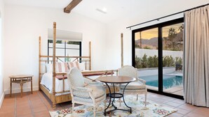 Un dormitorio con cama, dos sillas, una mesita y un amplio ventanal con vistas a una piscina y montañas.
