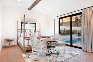 A bedroom with a bed, two chairs, a small table, and a large window with a view of a pool and mountains.