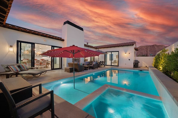 A modern outdoor pool area with lounge chairs and umbrellas, surrounded by a white building with large glass doors.