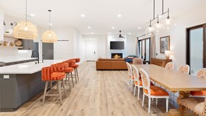 Una cocina moderna con una isla central, taburetes naranjas, un comedor con mesa de madera y sillas blancas, y una chimenea al fondo.