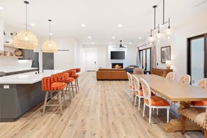 A modern kitchen with a central island, orange bar stools, a dining area with a wooden table and white chairs, and a fireplace in the background.