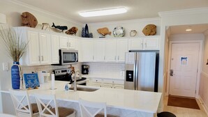 Een moderne keuken met witte kasten, een roestvrijstalen koelkast, een magnetron en een koffiezetapparaat. Er staan decoratieve items op het aanrecht en een afvalbak bij de deur.
