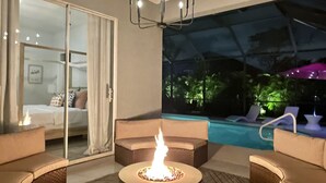 A modern indoor pool area with a fire pit, lounge chairs, and a glass door leading to a bedroom. - Beach + Heated Pool + Game Room + 2 King Suites (Naples)
