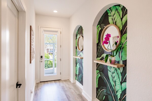 Un pasillo con una puerta blanca, piso de madera y un muro con estampado de hojas tropicales.
