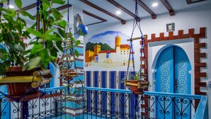 Un balcón con barandillas azules, plantas colgantes y un mural de un pueblo costero.
