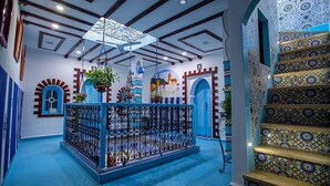 Una habitación con piso azul, un ventilador de techo decorativo, una escalera con diseño y una puerta azul con arco blanco.