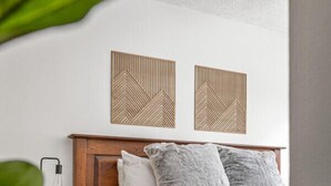 A neatly made bed with a wooden headboard, two wall-mounted geometric decor pieces, a bedside lamp, and a white rug. - Kauhoolio | Toby | Pool | Hot Tub | Sauna (Panorama)