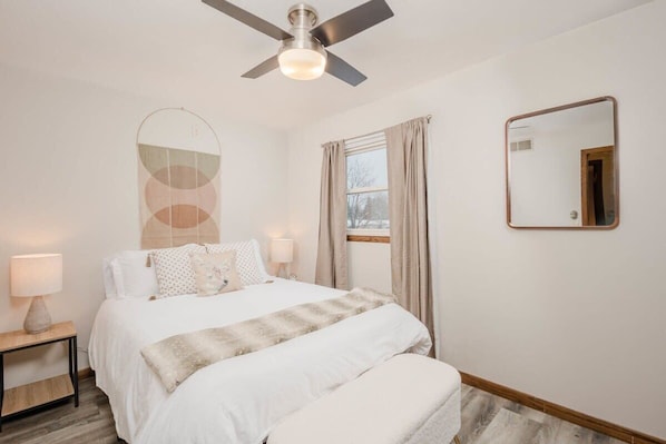 A bedroom with a bed, bedside tables, a ceiling fan, a window with curtains, and a mirror on the wall.