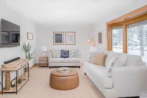 A living room with a sofa, ottoman, side tables, a TV, and framed pictures on the wall.