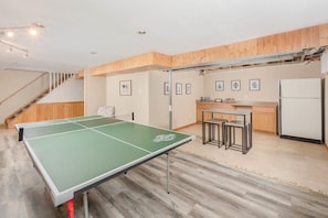A room with a ping pong table, wooden flooring, a kitchen area with a refrigerator, and a staircase.