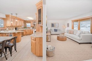 A modern kitchen with wooden cabinets and a dining area, a living room with a sofa and a coffee table, and large windows with a view of a snowy landscape. - Amazing Family Retreat w/ Fenced Yard & BBQ! (Stevens Point)