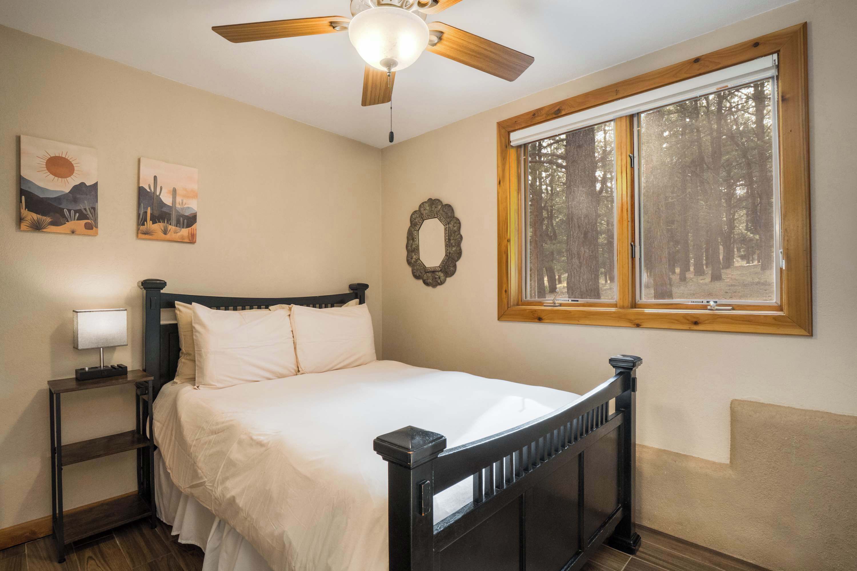 A bedroom with a bed, a nightstand, a ceiling fan, a window with a view of trees, and two paintings on the wall.