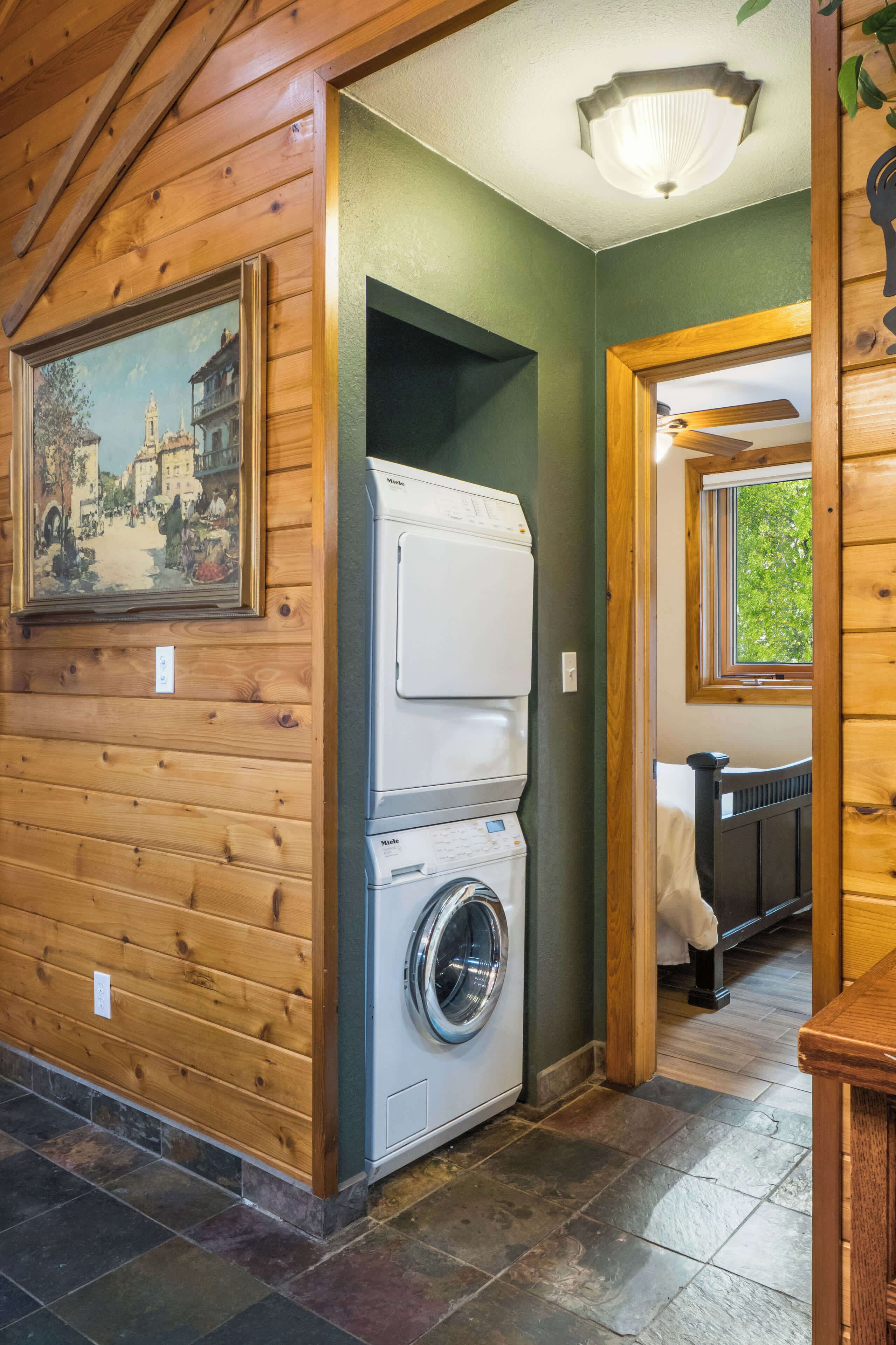 A laundry area with a stacked washer and dryer, a painting on the wall, and a window with a view.