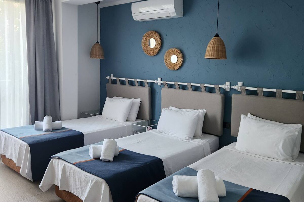 Een hotelkamer met vier bedden, elk voorzien van witte lakens en blauwe accenten, een grote spiegel en een plafondventilator.
