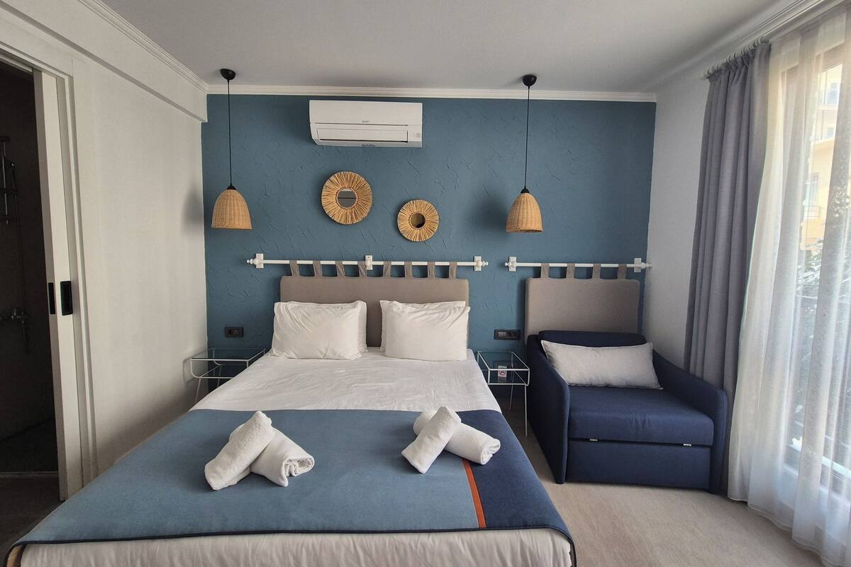 Een slaapkamer met een bed, nachtkastjes, een stoel, een airconditioning en een raam met gordijnen.