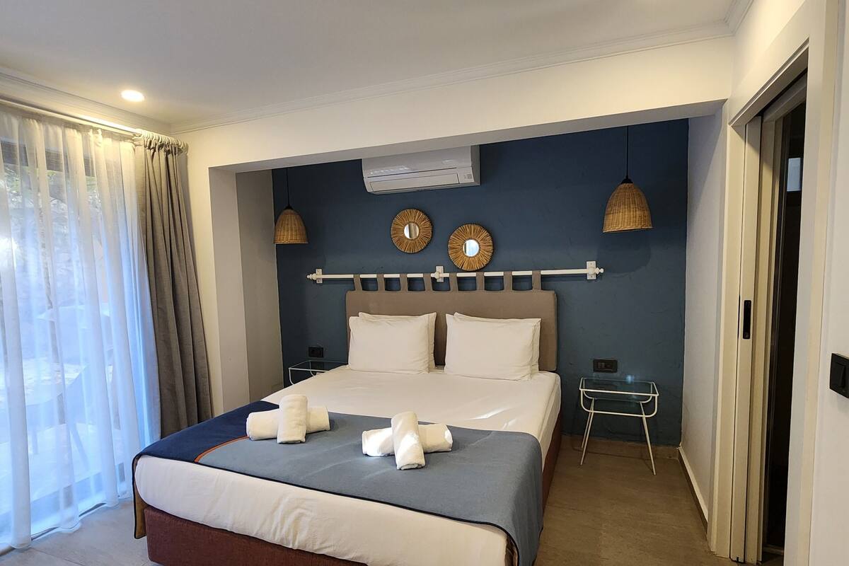 Een keurig opgemaakt bed met witte lakens en handdoeken, een nachtkastje met een lamp en een wandairconditioner.