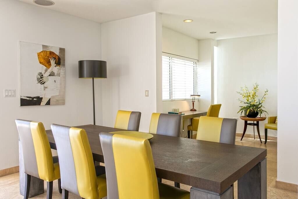 A modern dining area with a long table and chairs, a floor lamp, and a window with blinds.